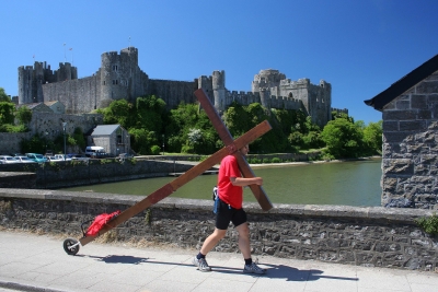 Counties evangelist Clive Cornish walking past Pembroke Castle with his cross. The magnificent medieval castle is viewed behind Clive as he crosses the West Wales river.